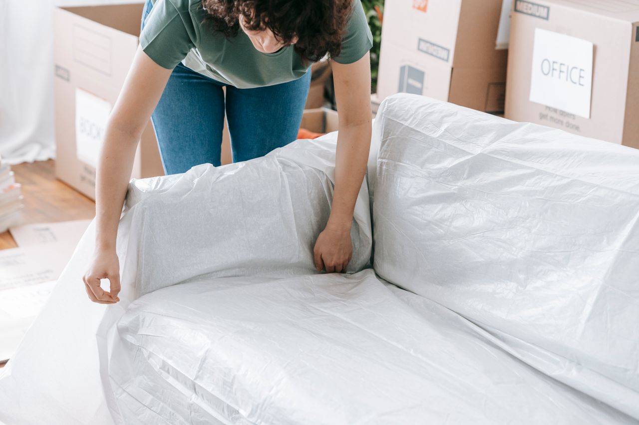 Une femme enveloppe avec soin un canapé dans un matériau de protection tout en remplissant des cartons dans une pièce lumineuse.
